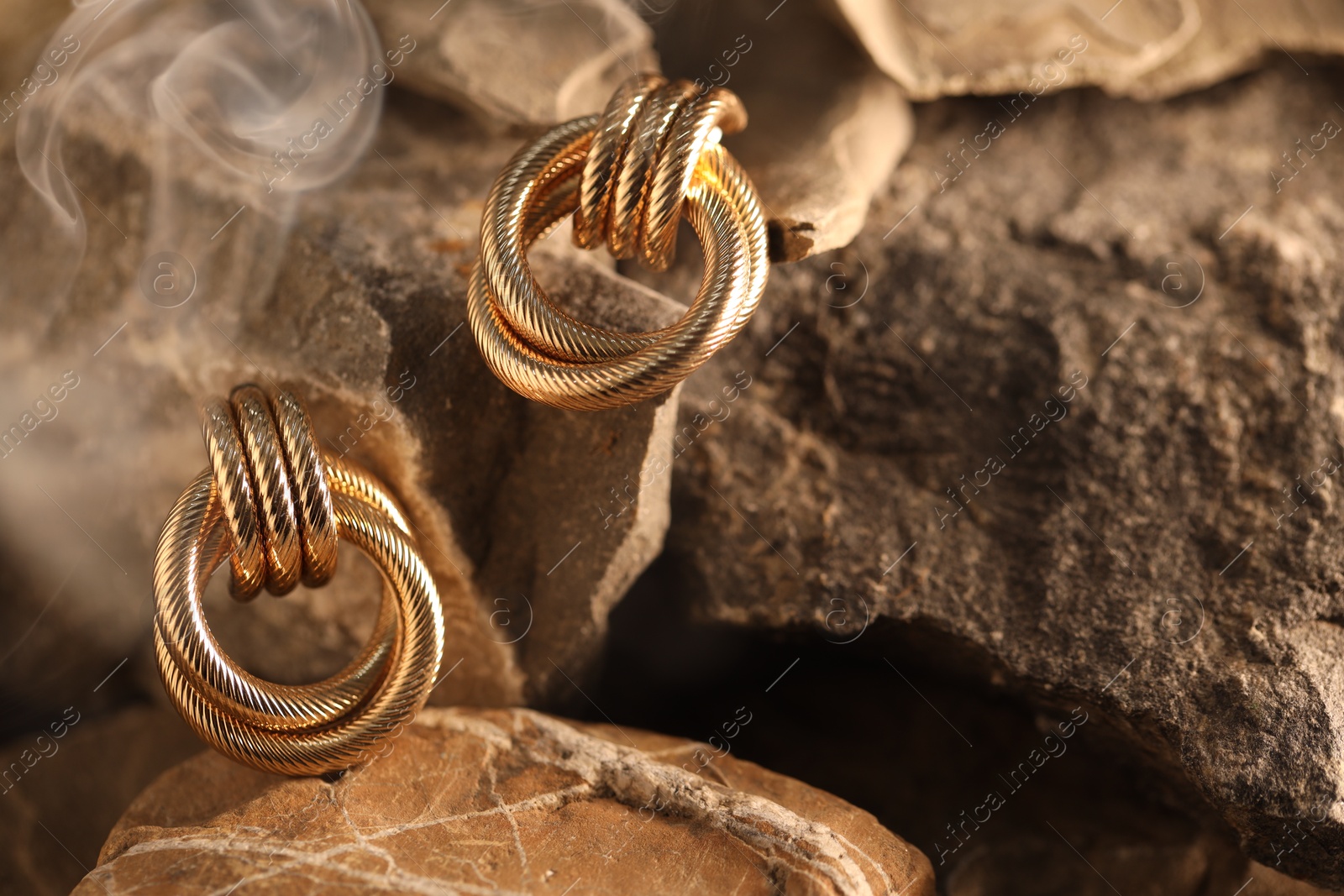Photo of Stylish gold earrings in smoke on rocks, closeup
