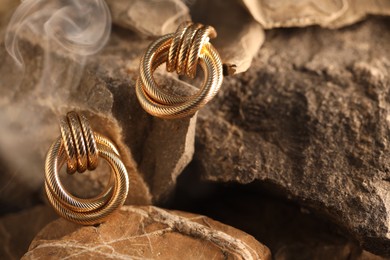 Photo of Stylish gold earrings in smoke on rocks, closeup