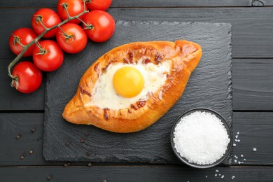 Photo of Tasty khachapuri with cheese, egg, tomatoes and spices on black wooden table, flat lay