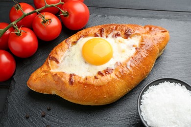 Photo of Tasty khachapuri with cheese, egg, tomatoes and spices on black wooden table, closeup