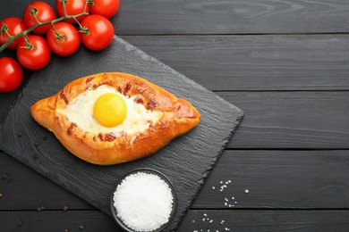 Photo of Tasty khachapuri with cheese, egg, tomatoes and spices on black wooden table, flat lay. Space for text