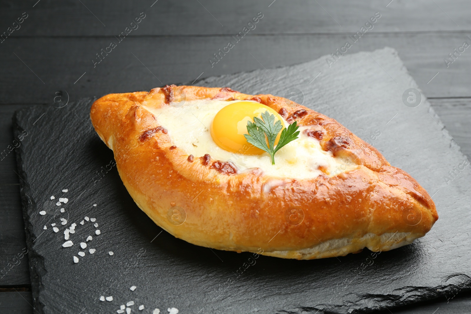 Photo of Tasty khachapuri with cheese, egg, salt and parsley on black table, closeup
