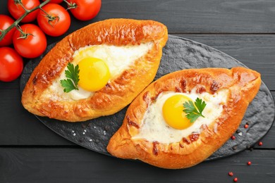 Photo of Tasty khachapuri with cheese, eggs, tomatoes and spices on black wooden table, flat lay