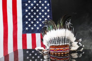 Photo of Traditional warbonnet (Native American chief headdress) in smoke on black mirror surface against flag of USA