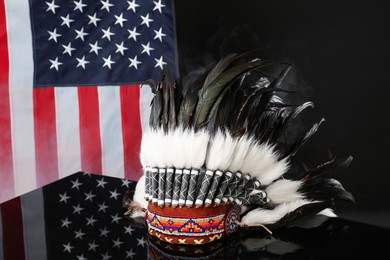 Photo of Traditional warbonnet (Native American chief headdress) in smoke on black mirror surface against flag of USA