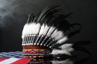 Photo of Traditional warbonnet (Native American chief headdress) and flag of USA in smoke on black mirror surface