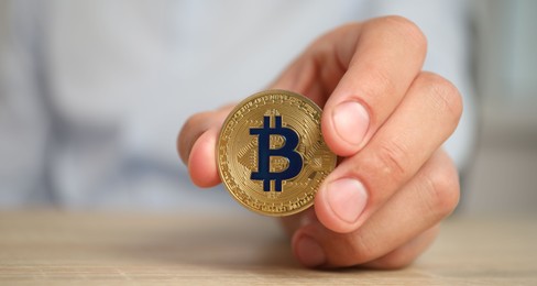 Photo of Cryptocurrency. Man with golden bitcoin at wooden table against blurred background, closeup. Space for text