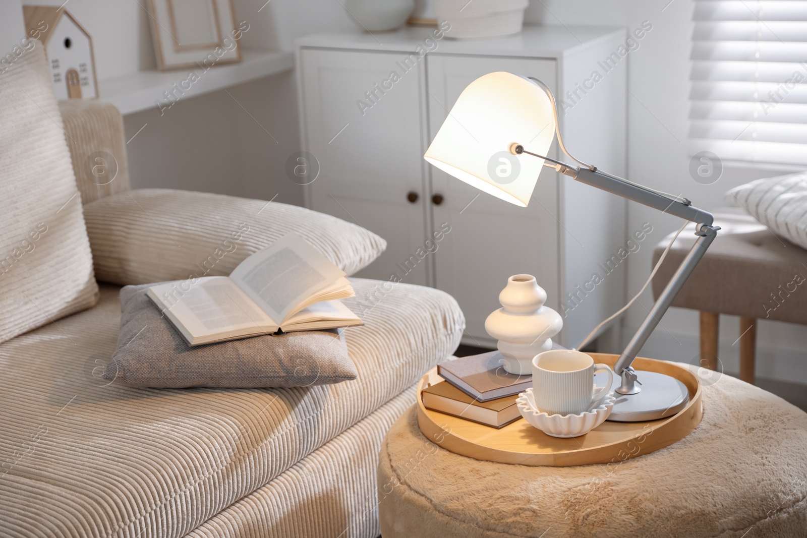 Photo of Stylish lamp, vase, books and cup on pouf near sofa indoors