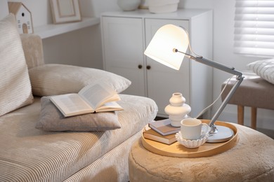 Photo of Stylish lamp, vase, books and cup on pouf near sofa indoors