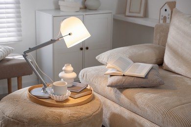 Photo of Stylish lamp, vase, books and cup on pouf near sofa indoors