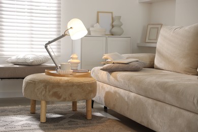 Photo of Stylish lamp, vase, books and cup on pouf near sofa indoors