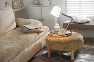 Photo of Stylish lamp, vase, books and cup on pouf near sofa indoors