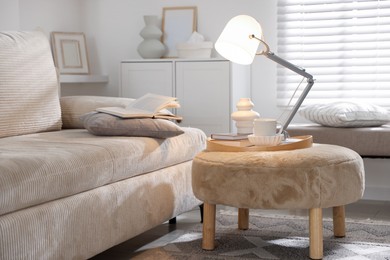 Photo of Stylish lamp, vase, books and cup on pouf near sofa indoors