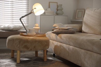 Photo of Stylish lamp, vase, books and cup on pouf near sofa indoors