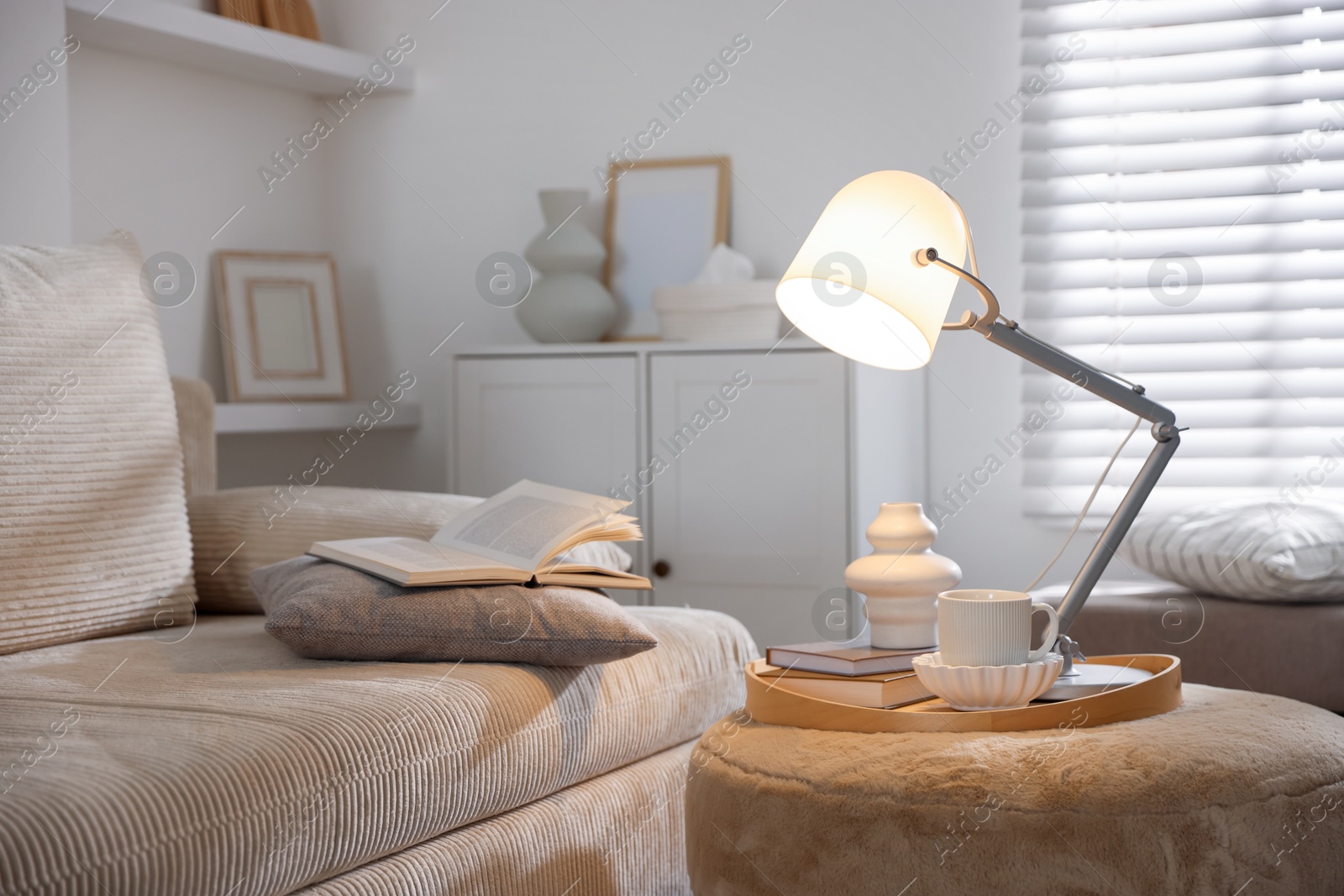 Photo of Stylish lamp, vase, books and cup on pouf near sofa indoors