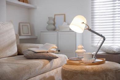 Photo of Stylish lamp, vase, books and cup on pouf near sofa indoors