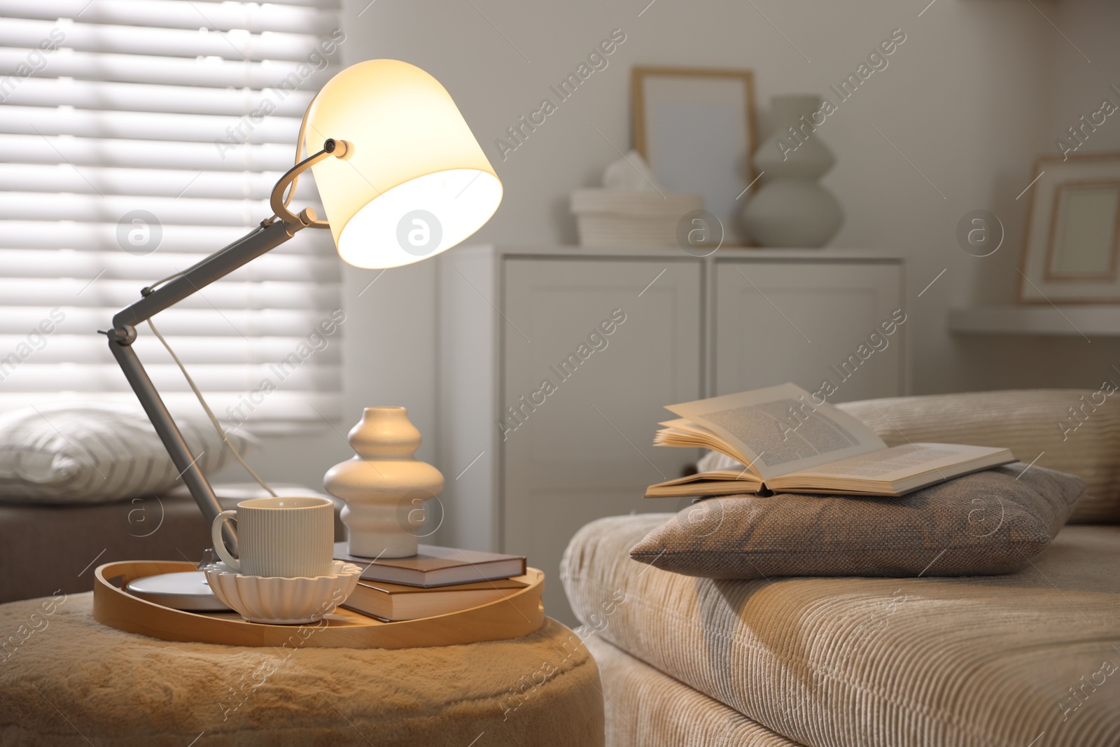Photo of Stylish lamp, vase, books and cup on pouf near sofa indoors