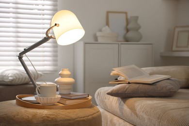 Photo of Stylish lamp, vase, books and cup on pouf near sofa indoors