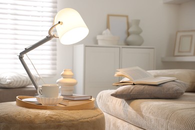 Photo of Stylish lamp, vase, books and cup on pouf near sofa indoors