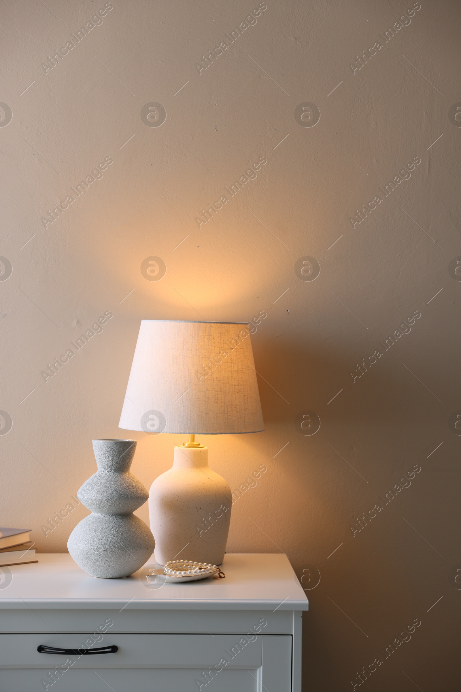 Photo of Stylish lamp, vase, pearl bracelet and books on white dresser near beige wall indoors