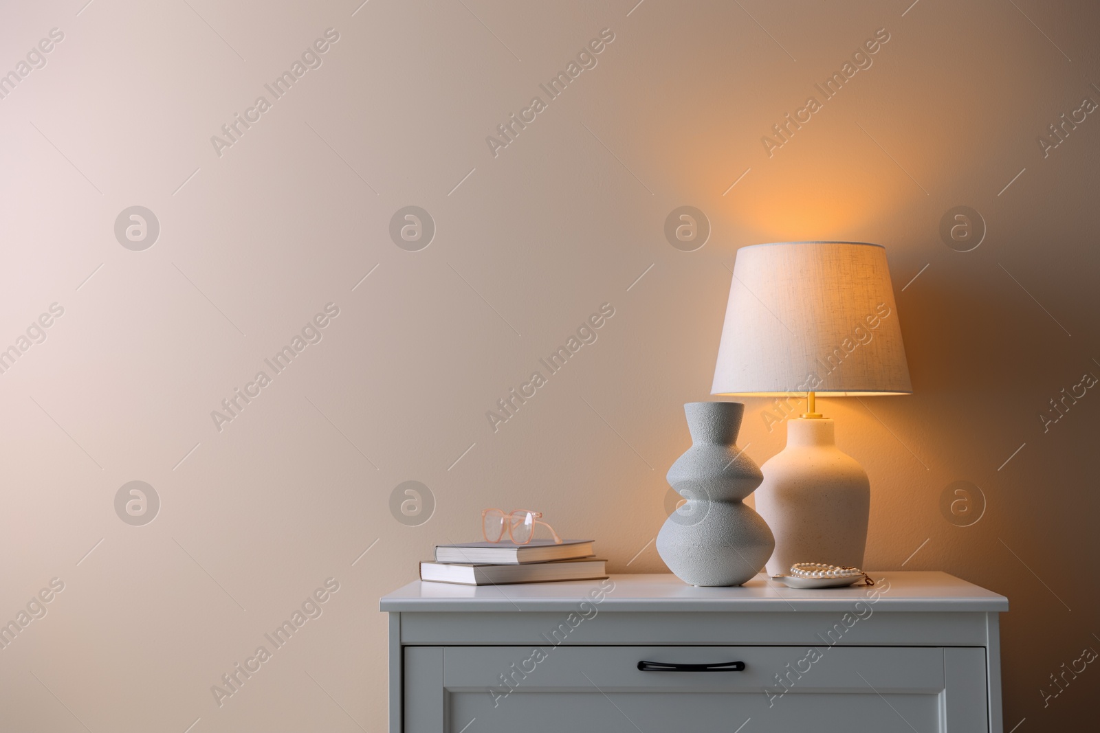 Photo of Stylish lamp, vase, pearl bracelet, books and glasses on white dresser near beige wall indoors, space for text