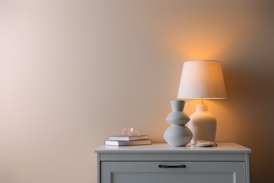 Photo of Stylish lamp, vase, pearl bracelet, books and glasses on white dresser near beige wall indoors, space for text
