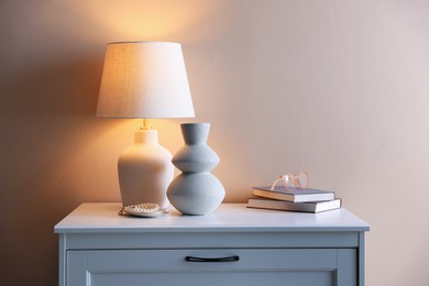 Photo of Stylish lamp, vase, pearl bracelet, books and glasses on white dresser near beige wall indoors