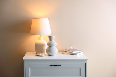 Photo of Stylish lamp, vase, pearl bracelet, books and glasses on white dresser near beige wall indoors