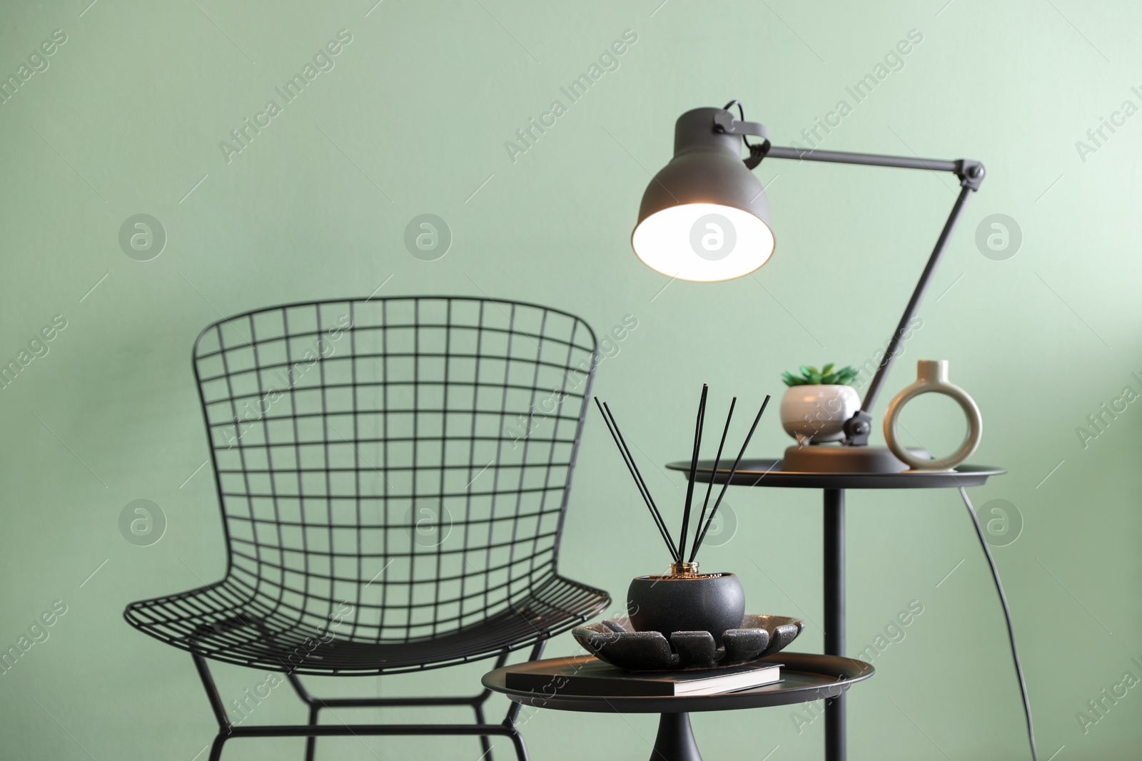 Photo of Stylish lamp, decor and reed diffuser on nesting tables near armchair against green wall indoors