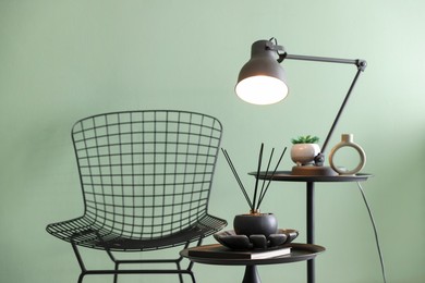 Photo of Stylish lamp, decor and reed diffuser on nesting tables near armchair against green wall indoors