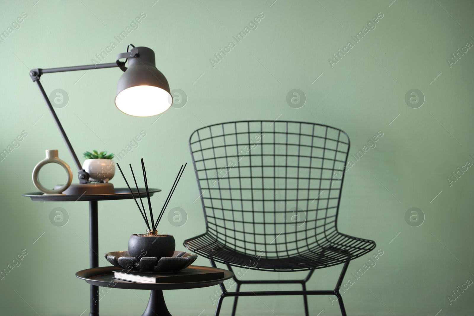 Photo of Stylish lamp, decor and reed diffuser on nesting tables near armchair against green wall indoors