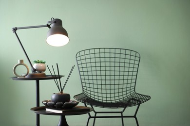 Photo of Stylish lamp, decor and reed diffuser on nesting tables near armchair against green wall indoors