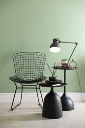 Photo of Stylish lamp, decor and reed diffuser on nesting tables near armchair against green wall indoors