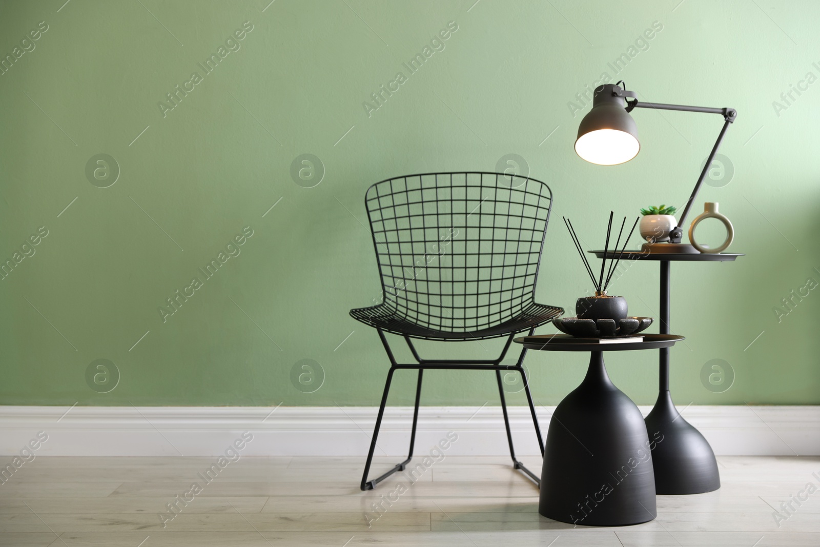 Photo of Stylish lamp, decor and reed diffuser on nesting tables near armchair against green wall indoors, space for text