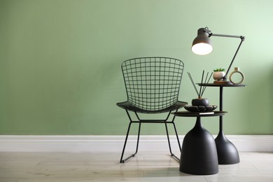 Photo of Stylish lamp, decor and reed diffuser on nesting tables near armchair against green wall indoors, space for text