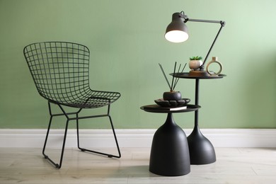 Photo of Stylish lamp, decor and reed diffuser on nesting tables near armchair against green wall indoors