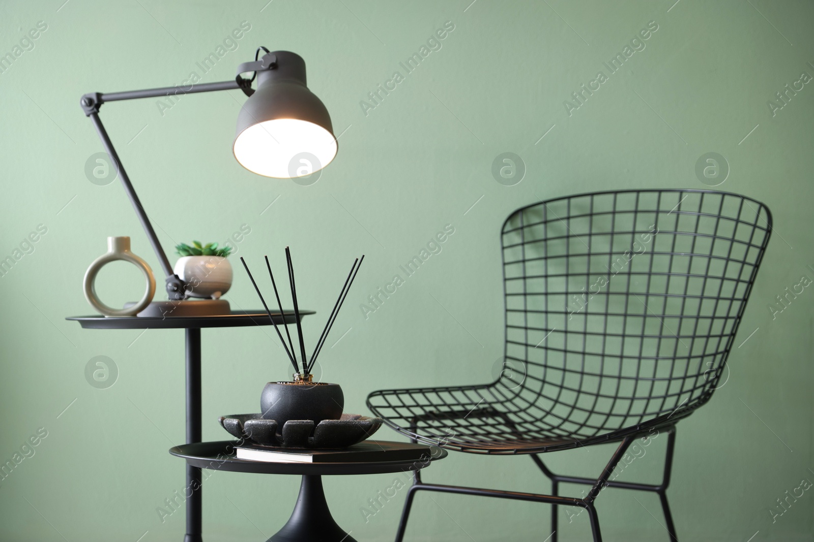 Photo of Stylish lamp, decor and reed diffuser on nesting tables near armchair against green wall indoors