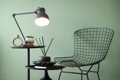 Photo of Stylish lamp, decor and reed diffuser on nesting tables near armchair against green wall indoors