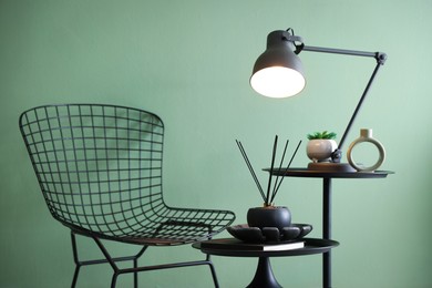Photo of Stylish lamp, decor and reed diffuser on nesting tables near armchair against green wall indoors