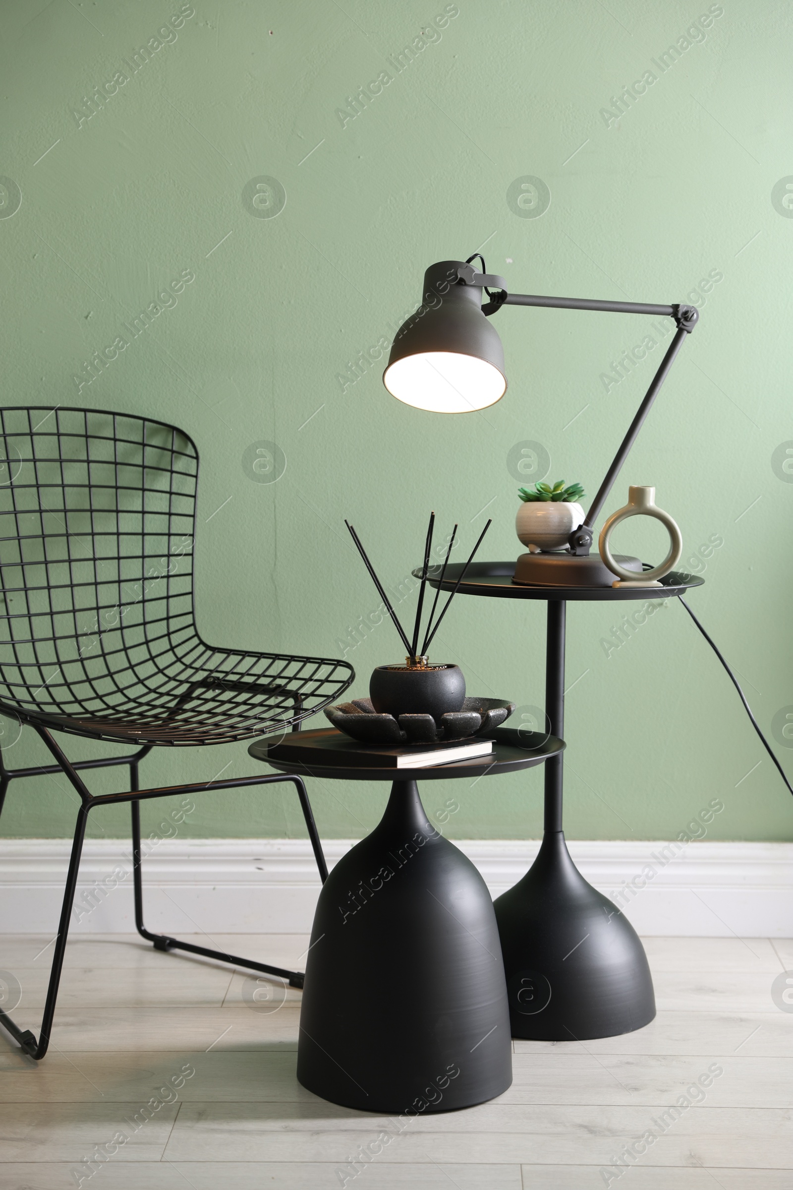 Photo of Stylish lamp, decor and reed diffuser on nesting tables near armchair against green wall indoors