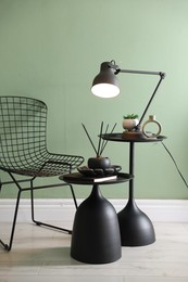 Photo of Stylish lamp, decor and reed diffuser on nesting tables near armchair against green wall indoors