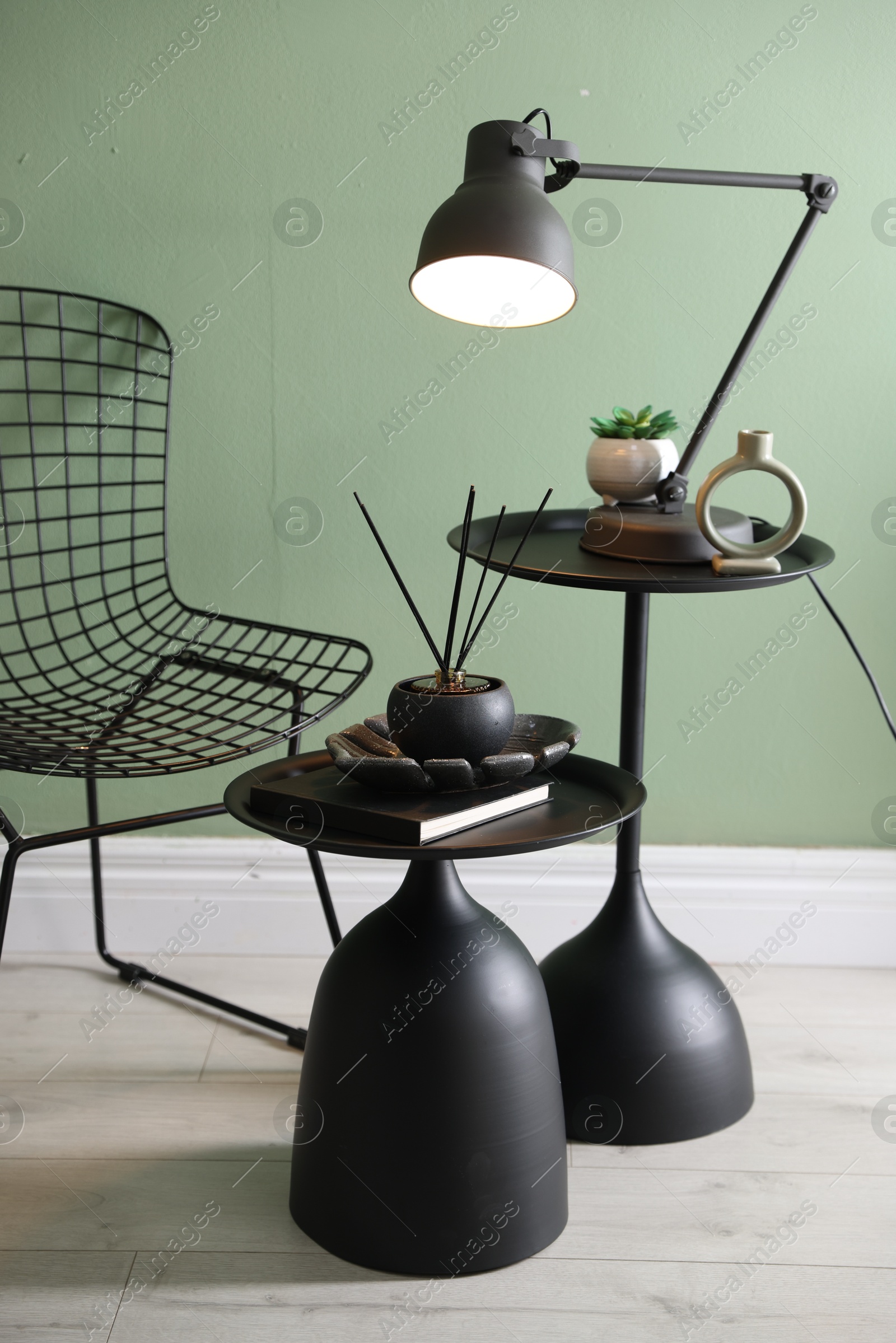 Photo of Stylish lamp, decor and reed diffuser on nesting tables near armchair against green wall indoors