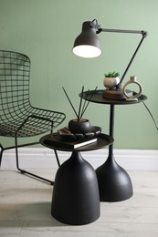 Photo of Stylish lamp, decor and reed diffuser on nesting tables near armchair against green wall indoors