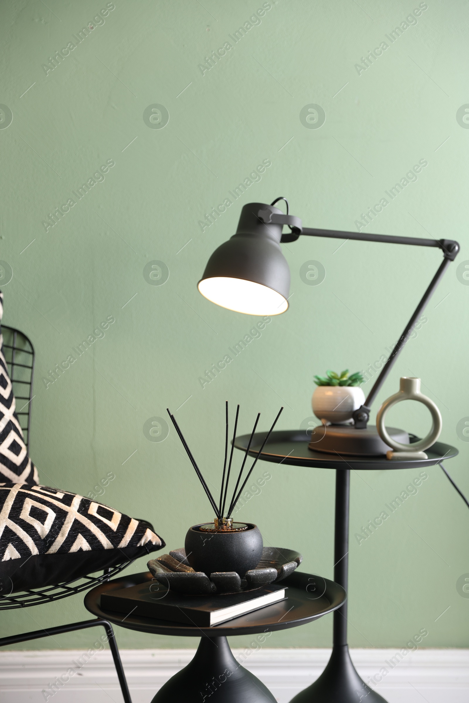 Photo of Stylish lamp, decor and reed diffuser on nesting tables near armchair against green wall indoors