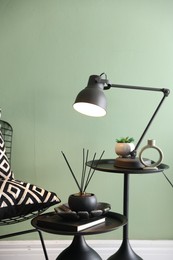 Photo of Stylish lamp, decor and reed diffuser on nesting tables near armchair against green wall indoors