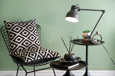 Photo of Stylish lamp, decor and reed diffuser on nesting tables near armchair against green wall indoors