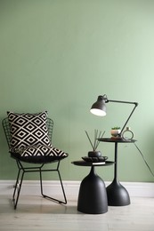 Photo of Stylish lamp, decor and reed diffuser on nesting tables near armchair against green wall indoors
