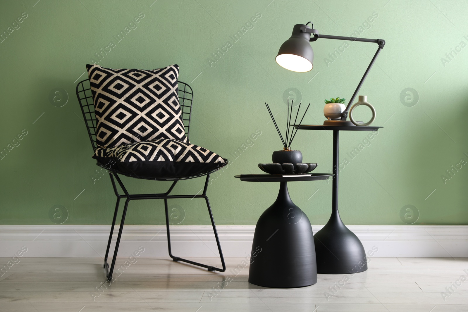 Photo of Stylish lamp, decor and reed diffuser on nesting tables near armchair against green wall indoors