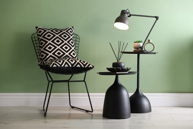 Photo of Stylish lamp, decor and reed diffuser on nesting tables near armchair against green wall indoors
