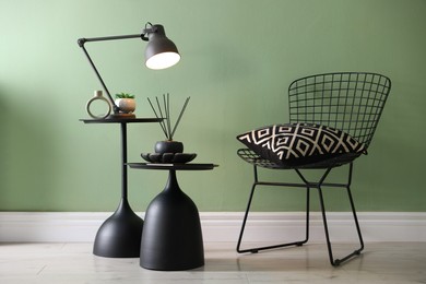 Photo of Stylish lamp, decor and reed diffuser on nesting tables near armchair against green wall indoors